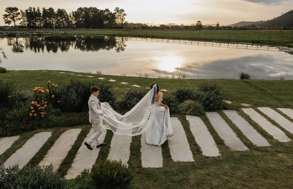 Couple walking across stepping stones beside a lake at sunset, the veil trailing